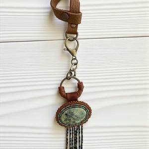 May include: A handcrafted keychain with a brown leather loop and a metal clasp. The pendant features an oval, green stone surrounded by beaded detailing and dangling beaded fringe. The keychain is designed with a mix of textures and colors.