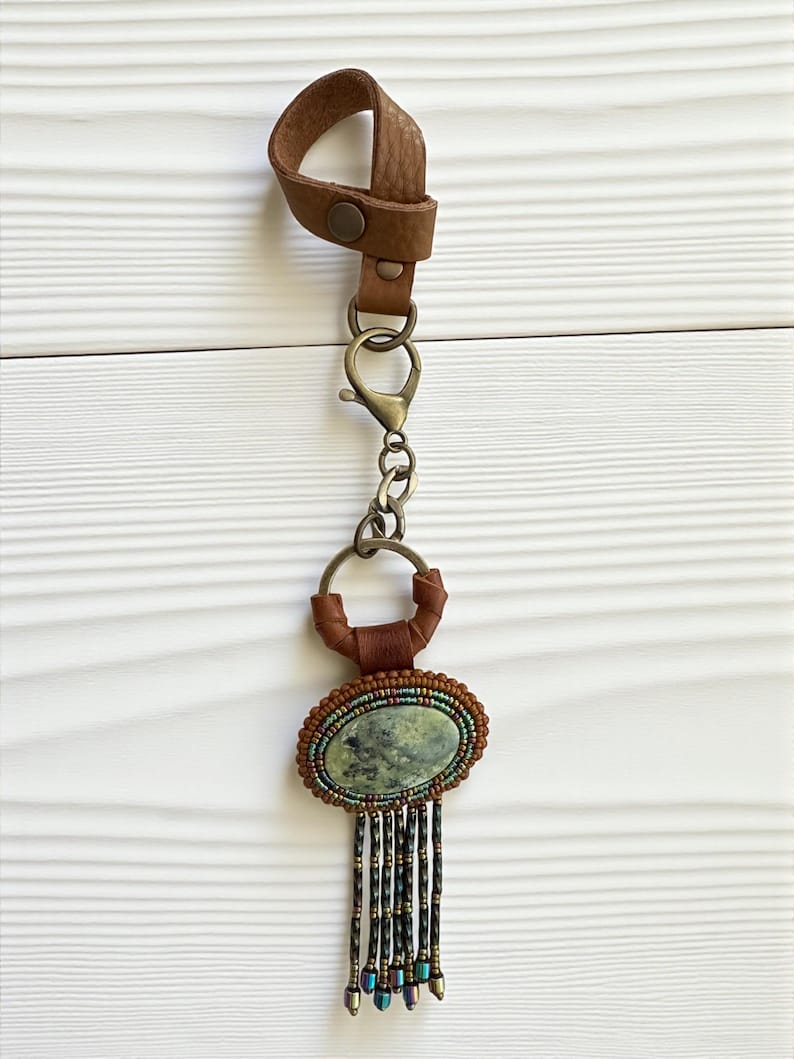 May include: A handcrafted keychain with a brown leather loop and a metal clasp. The pendant features an oval, green stone surrounded by beaded detailing and dangling beaded fringe. The keychain is designed with a mix of textures and colors.