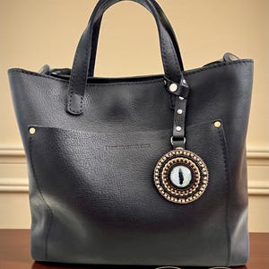 May include: Black leather tote bag with a decorative eye charm and a pair of sunglasses. The tote has two handles and a small pocket on the front. The charm is round with a beaded design and a blue eye in the center.