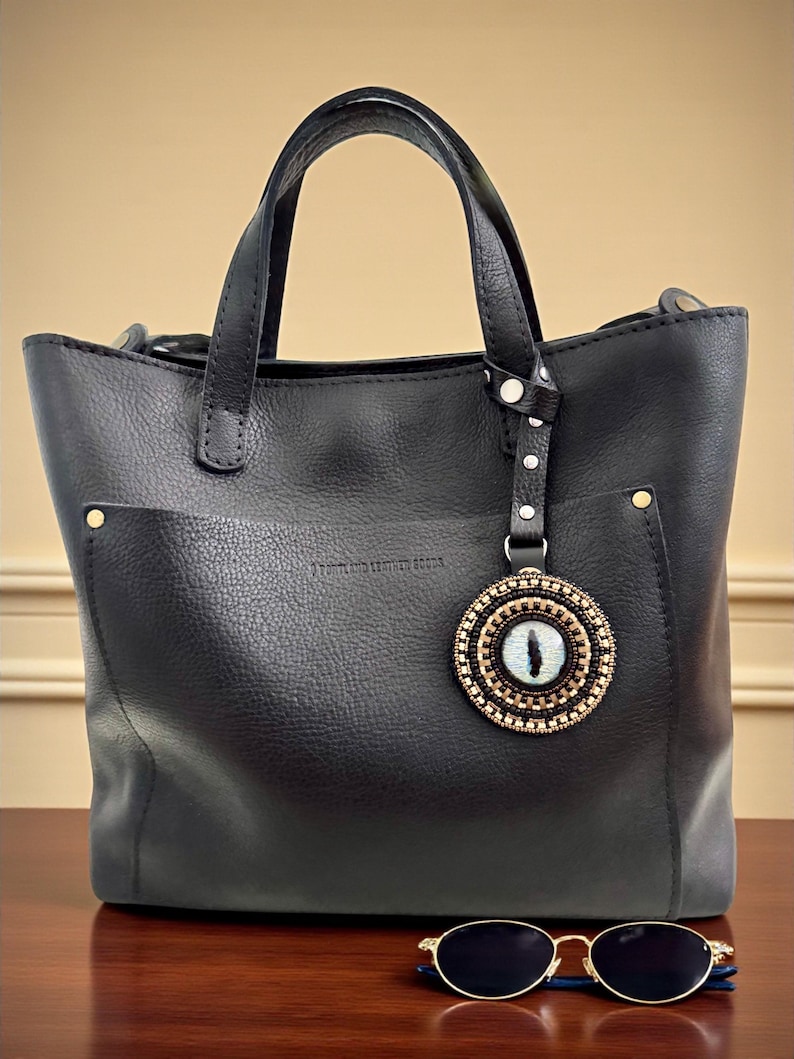 May include: Black leather tote bag with a decorative eye charm and a pair of sunglasses. The tote has two handles and a small pocket on the front. The charm is round with a beaded design and a blue eye in the center.