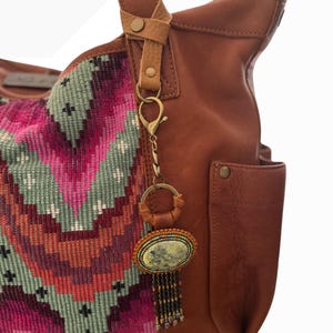 May include: A brown leather tote bag with a colorful woven fabric panel. A decorative keychain with an oval stone and beaded fringe hangs from the handle. The bag has a pocket and leather strap details.