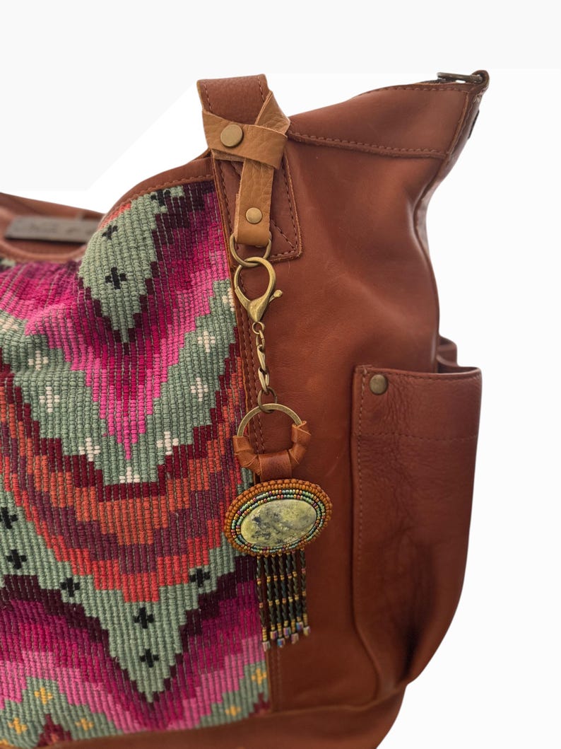 May include: A brown leather tote bag with a colorful woven fabric panel. A decorative keychain with an oval stone and beaded fringe hangs from the handle. The bag has a pocket and leather strap details.