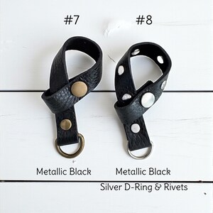May include: Two black leather accessories with metallic accents are displayed against a white wooden background. One has a gold-colored rivet and D-ring, while the other features silver rivets and a silver D-ring. The text reads "Metallic Black" and "Silver D-Ring & Rivets."