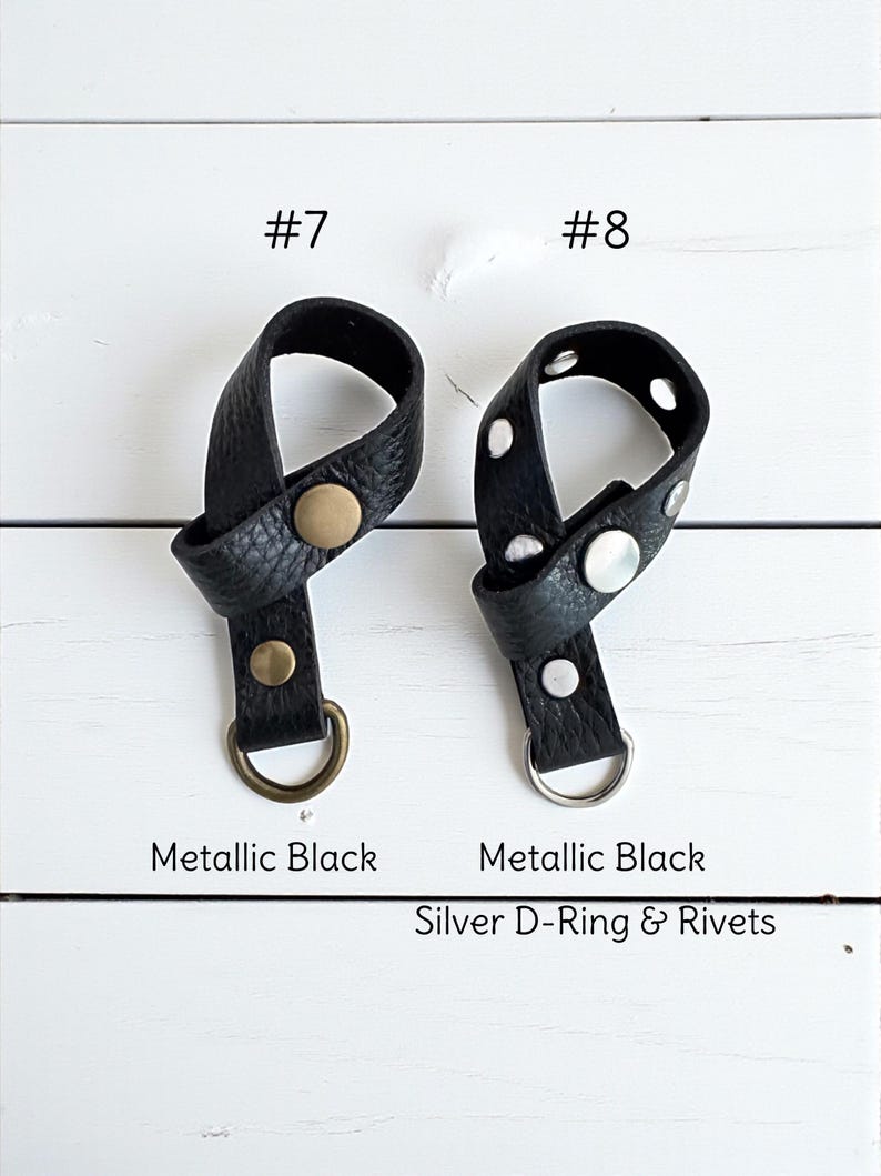 May include: Two black leather accessories with metallic accents are displayed against a white wooden background. One has a gold-colored rivet and D-ring, while the other features silver rivets and a silver D-ring. The text reads "Metallic Black" and "Silver D-Ring & Rivets."