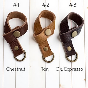 May include: Leather wrist straps in three colours: Chestnut, Tan, and Dk. Expresso. Each strap features a brass button and a metal D-ring. The straps are displayed against a white wooden background.