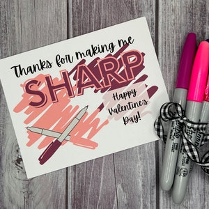 Puede incluir: Una tarjeta de San Valentín con el texto "Gracias por hacerme AGUDO" y un dibujo de dos marcadores. La tarjeta está junto a tres marcadores atados con una cinta blanca y negra.