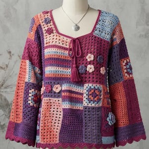 May include: A long-sleeved crochet sweater featuring a patchwork design in shades of purple, pink, orange, and light blue. The garment has a square neckline with a drawstring tie and tassel, accented with small three-dimensional crochet flowers.
