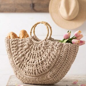Op de afbeelding: Een beige gehaakte handtas met een halfronde vorm en twee ronde bamboe handvatten. De tas is gevuld met twee knapperige broodjes aan de linkerkant en een boeketje lichtroze tulpen dat aan de rechterkant uit de tas steekt.