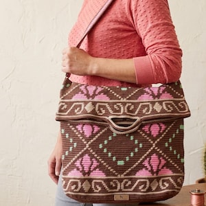 Könnte beinhalten: Eine braune Häkel-Umhängetasche mit einem geometrischen Muster in Pink und Hellgrün. Die Tasche hat einen Umschlag, einen breiten Schulterriemen und ein kleines rechteckiges Etikett mit dem Buchstaben N in der unteren Mitte der Vorderseite.