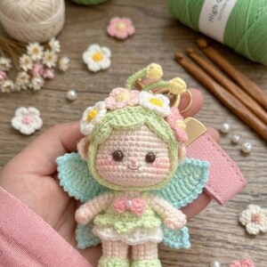 Könnte beinhalten: Ein Häkelpuppen-Schlüsselanhänger, der eine Figur mit einer hellgrünen Kapuze zeigt, die mit kleinen rosa und weißen Blumen verziert ist. Die Puppe hat hellblaue Flügel, eine rosa Schleife am Hals und ein hellrosa Kunstlederband an einem Goldring.