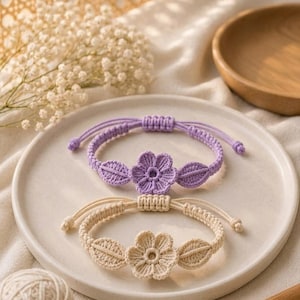 May include: Two handmade crochet bracelets with flower and leaf designs. One is lavender, the other cream. Both are on a white plate with yarn and a crochet hook.