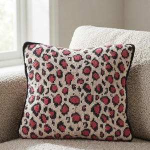 Könnte beinhalten: Ein quadratisches Strickkissen mit einem sich wiederholenden Leopardenmuster in dunklem Anthrazit und leuchtendem Pink auf cremefarbenem Hintergrund, das an den Außenkanten mit einem dunkelgrauen Rand versehen ist.