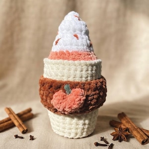 Könnte beinhalten: Ein plüschiger Häkelartikel in Form eines Kaffeebechers mit einer Sahnehaube. Der Becher ist weiß mit einem dunkelbraunen Mittelband, das eine kleine orangefarbene Kürbisapplikation aufweist. Die Oberseite ist weiß mit orangefarbenen Akzenten.
