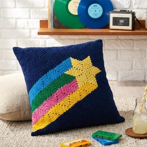 Könnte beinhalten: Ein quadratisches, dunkelblaues Häkelkissen mit einem großen gelben Stern und einem regenbogenfarbenen Schweif in Blau, Grün und Pink. Das Kissen weist ein strukturiertes, sich wiederholendes geometrisches Muster auf dem dunkelblauen Grund auf.