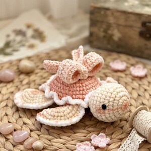 May include: A crocheted turtle toy in soft pastel pink and cream yarn. The turtle features a rounded shell with a ruffled edge, a large decorative bow on top, and a round head with small black bead eyes. The yarn has a plush, velvet-like texture.