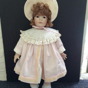 Puede incluir: Una muñeca de porcelana que lleva un vestido rosa con ribete de encaje y un sombrero blanco con flores rosas. La muñeca tiene el pelo rojo y los ojos azules.