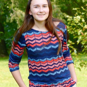 May include: A young person with long brown hair wearing a blue, pink, and red chevron patterned long-sleeved shirt and khaki pants.