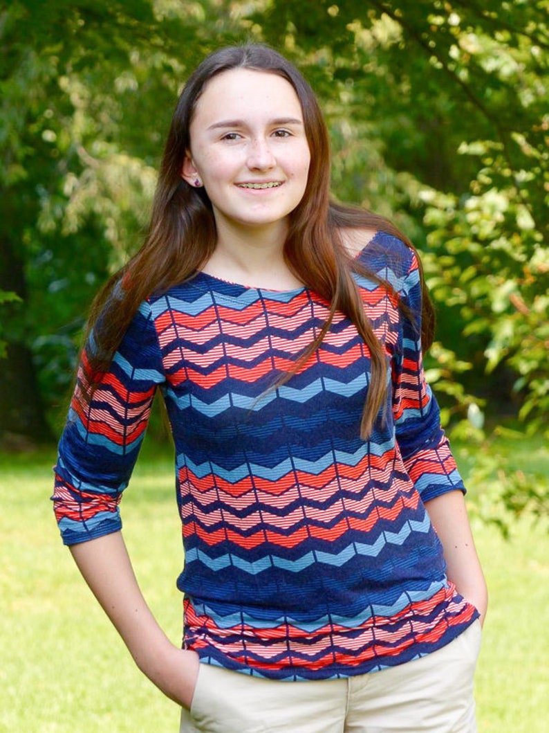 May include: A young person with long brown hair wearing a blue, pink, and red chevron patterned long-sleeved shirt and khaki pants.