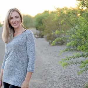 May include: A woman wearing a gray, long-sleeved shirt with a scoop neck. She is standing in a desert landscape with green bushes in the background.