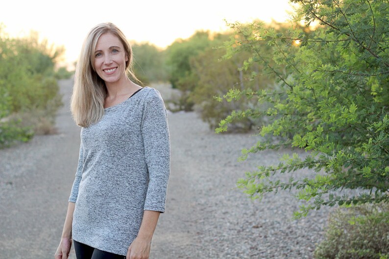 May include: A woman wearing a gray, long-sleeved shirt with a scoop neck. She is standing in a desert landscape with green bushes in the background.