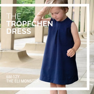 Può includere: Una giovane persona indossa un abito blu navy senza maniche con un davanti plissettato. L'abito ha una scollatura rotonda ed è etichettato "THE TRÖPFCHEN DRESS 6M-12Y THE ELI MONSTER".