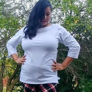 May include: A woman wearing a white, 3/4 sleeve, boat neck top with a plaid pattern pants.