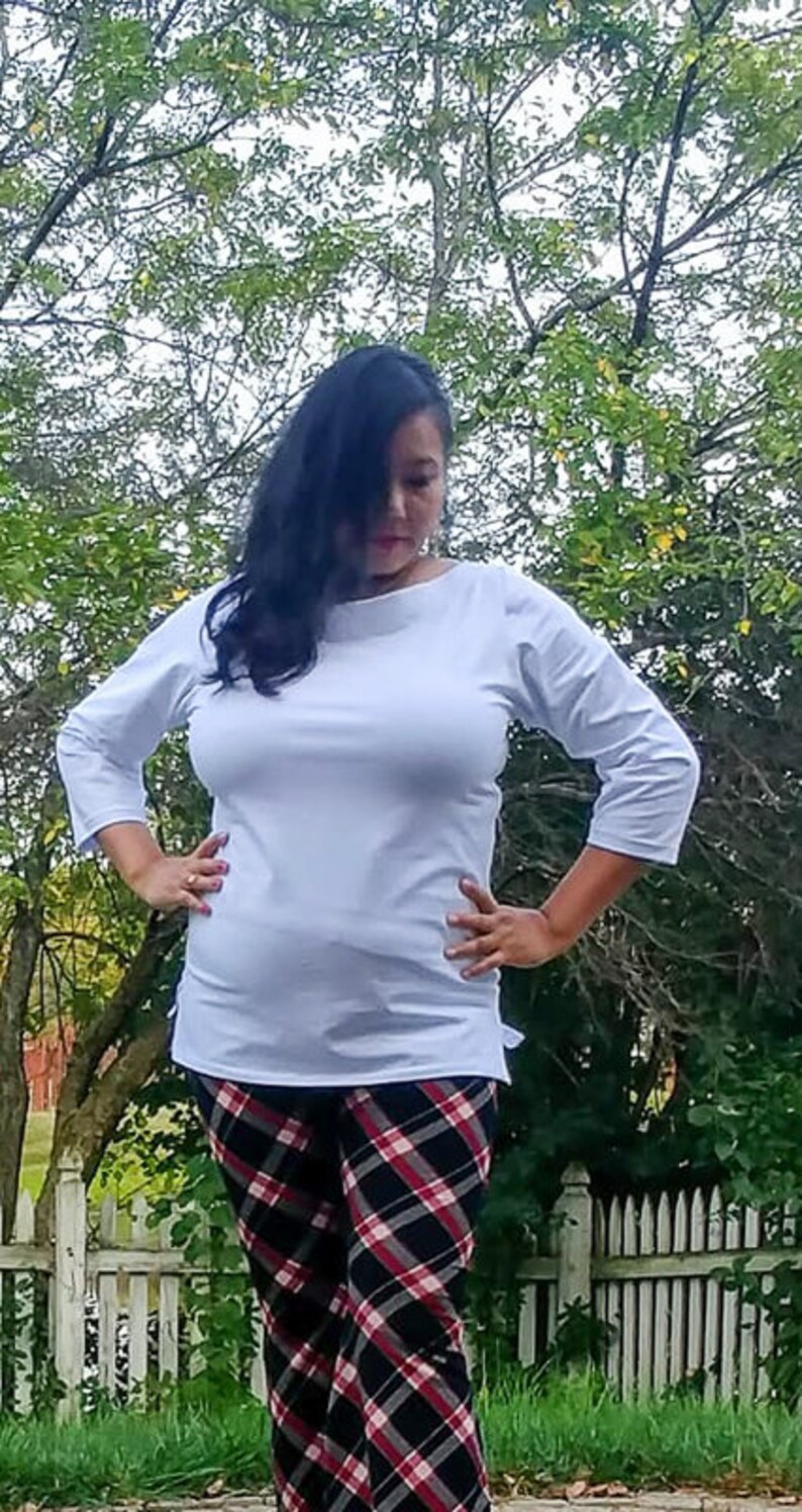 May include: A woman wearing a white, 3/4 sleeve, boat neck top with a plaid pattern pants.