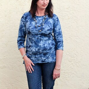 May include: A woman wearing a blue tie-dye shirt with a silver necklace and a gold bracelet. She is wearing blue jeans.