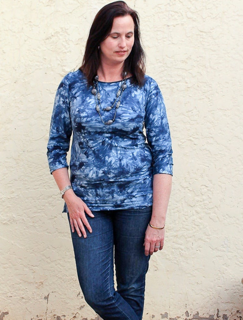 May include: A woman wearing a blue tie-dye shirt with a silver necklace and a gold bracelet. She is wearing blue jeans.