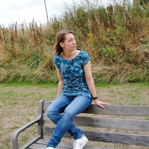 May include: A woman wearing a teal blue and white patterned shirt and blue jeans sits on a wooden bench. She is wearing white sneakers with silver accents.