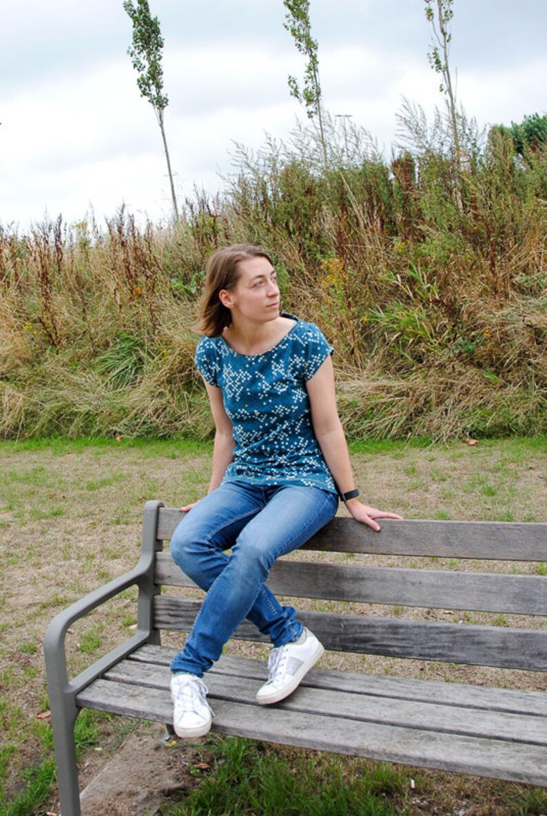 May include: A woman wearing a teal blue and white patterned shirt and blue jeans sits on a wooden bench. She is wearing white sneakers with silver accents.