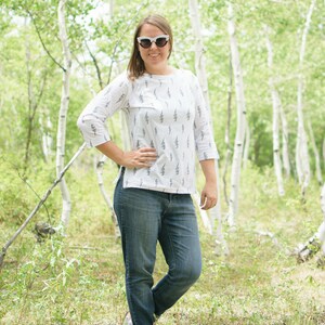 May include: A woman wearing white sunglasses and a white shirt with a lightning bolt pattern stands in a forest. She is wearing blue jeans and brown sandals.