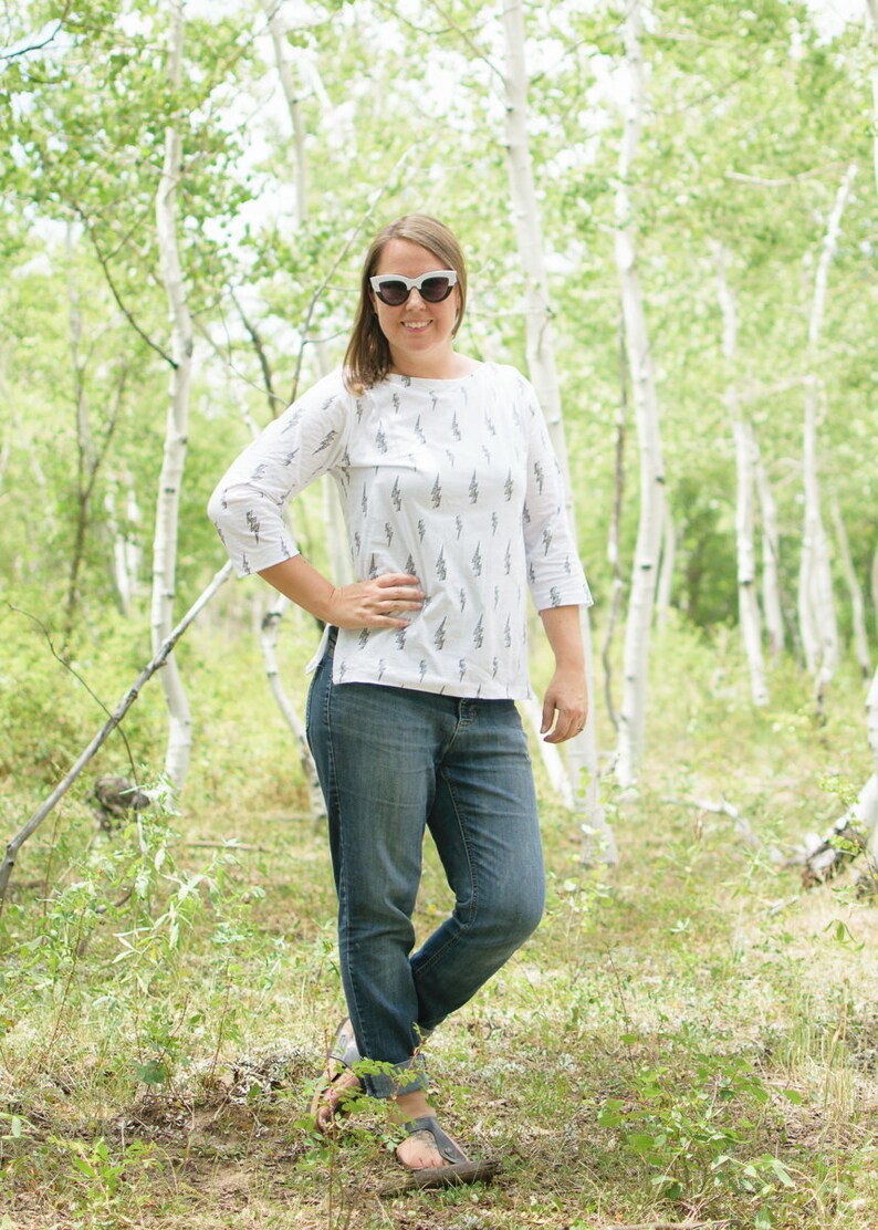 May include: A woman wearing white sunglasses and a white shirt with a lightning bolt pattern stands in a forest. She is wearing blue jeans and brown sandals.