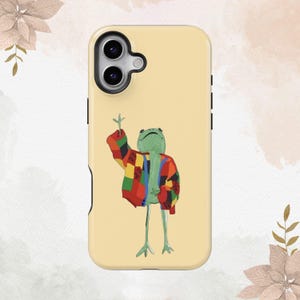 May include: A yellow phone case with a green frog wearing a colorful sweater. The frog is standing on two legs and has its arms raised.