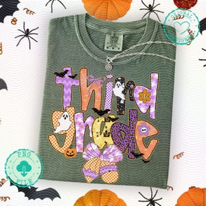 K&ouml;nnte beinhalten: Ein salbeigr&uuml;nes T-Shirt mit dem Schriftzug "Third Grade" in einem farbenfrohen Halloween-Design. Das Design umfasst Geister, Flederm&auml;use, K&uuml;rbisse und eine gro&szlig;e Schleife. Eine silberne Halskette mit Anh&auml;nger liegt auf dem Shirt.
