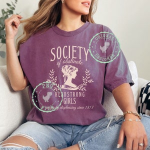 Puede incluir: Una camiseta morada con el texto "Society of obstinate headstrong girls" y una silueta de una mujer. El texto también incluye "displeasing since 1813".