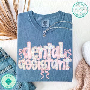 K&ouml;nnte beinhalten: Blaues Comfort Colors T-Shirt mit den Worten "dental assistant" in einem Blumen- und Vichy-Muster. Das Shirt hat rosa Schleifen und eine silberne Halskette. Das Shirt ist auf einer wei&szlig;en Oberfl&auml;che gefaltet.