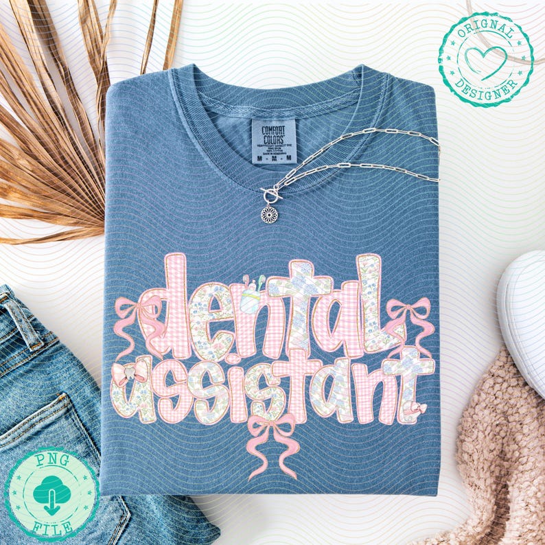 K&ouml;nnte beinhalten: Blaues Comfort Colors T-Shirt mit den Worten "dental assistant" in einem Blumen- und Vichy-Muster. Das Shirt hat rosa Schleifen und eine silberne Halskette. Das Shirt ist auf einer wei&szlig;en Oberfl&auml;che gefaltet.