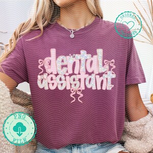 K&ouml;nnte beinhalten: Ein weinrotes T-Shirt mit dem Schriftzug "dental assistant" in einer dekorativen Schriftart. Der Text ist mit floralen und Vichy-Mustern sowie rosa Schleifen verziert. Das Shirt hat kurze &Auml;rmel und eine lockere Passform.