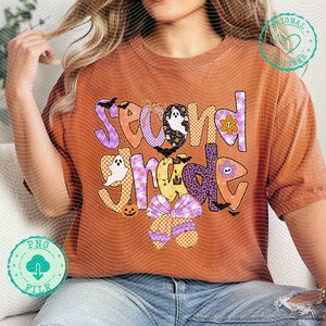 K&ouml;nnte beinhalten: Gebrannt orangefarbenes T-Shirt mit einem farbenfrohen "Second Grade"-Aufdruck. Das Design zeigt Geister, Flederm&auml;use, einen K&uuml;rbis und eine Schleife. Der Text ist in verschiedenen Mustern und Farben gehalten, darunter Lila, Orange und Gelb. Ein lustiges, festliches Shirt f&uuml;r das Schuljahr.
