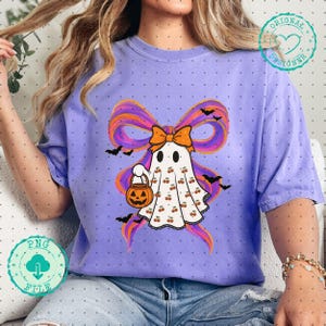 May include: A lilac-colored t-shirt featuring a Halloween ghost design. The ghost is adorned with cherries and holds a jack-o'-lantern. A large bow with orange and purple accents and black bats complete the design.