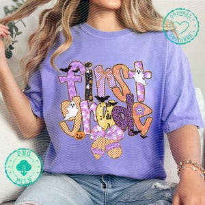 K&ouml;nnte beinhalten: Ein helllila T-Shirt mit dem Schriftzug "First Grade" in einem farbenfrohen Halloween-Design. Das Design umfasst Geister, Flederm&auml;use, K&uuml;rbisse und eine gro&szlig;e Schleife. Das Shirt ist ein bequemer, l&auml;ssiger Stil.