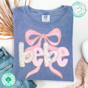 May include: A blue Comfort Colors t-shirt featuring a pink bow with gold glitter accents and the word "bebe" in a floral and gingham pattern. The shirt has a crew neck and short sleeves. A PNG file download is available.