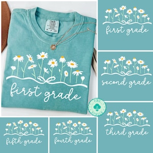 Può includere: T-shirt color verde acqua con un motivo floreale a margherite e la scritta "first grade" in bianco. Una collana d'oro è appoggiata sulla maglietta. Grafiche aggiuntive mostrano disegni simili per la seconda, terza, quarta e quinta elementare.