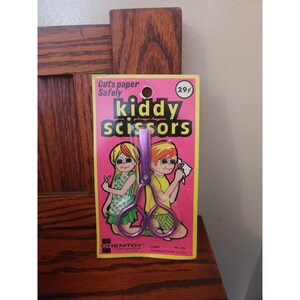May include: A packet of vintage "kiddy scissors" with a pink and yellow background. The package features illustrations of children and the text "Guts paper safely". The scissors are purple.