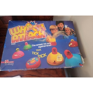 May include: A vintage Whack Attack game box with a blue background and colorful game pieces. The box features the game's title in large, bold letters and images of people playing. The game pieces are various colors, including pink, green, yellow, and blue.