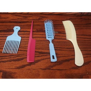 May include: Four hair combs of different styles and colors are displayed on a wooden surface. There is a light blue pick comb, a pink comb, a light blue comb with a handle, and a cream-colored comb.