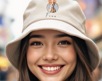Puede incluir: Una mujer con un sombrero de cubo beige con una ilustración de un perro y el nombre "Finn" en él. Ella está sonriendo y tiene el pelo largo y castaño.