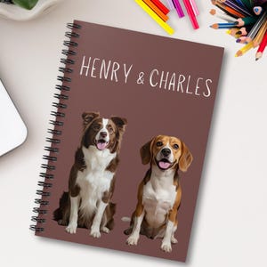 Puede incluir: Una libreta espiral marrón con los nombres "Henry & Charles" impresos en blanco. La libreta presenta ilustraciones de dos perros, un border collie marrón y blanco y un beagle, sentados sobre un fondo marrón.