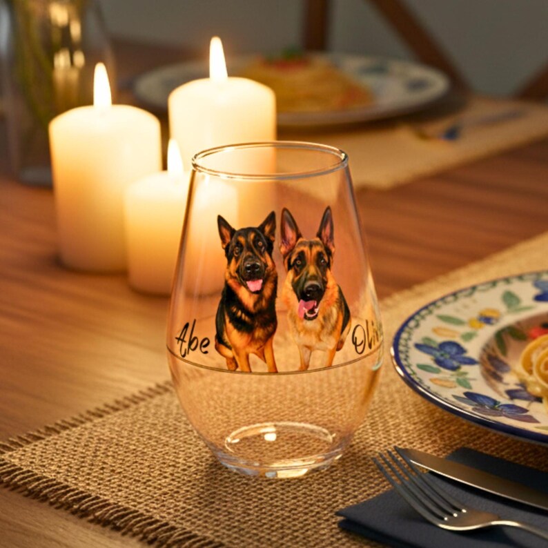 May include: A clear stemless wine glass with a custom design of two German Shepherds. The dogs have black and tan fur. The names "Abe" and "Olive" are written on the glass. The glass is on a burlap placemat. Candles and a plate are in the background.